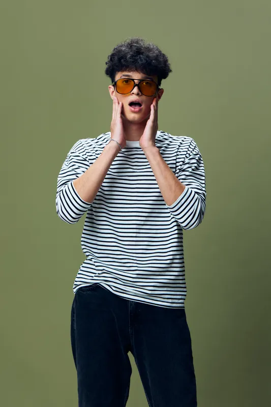surprised young man wearing striped shirt and orange sunglasses standing with hands on face against green background, expressing shock and amazement