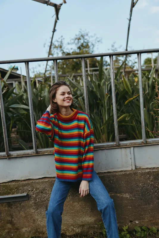 Woman in colorful sweater with a warm smile, casual sitting pose outdoors in a portrait wearing jeans and knitwear by a fence, relaxed fashion and cozy autumn vibe.