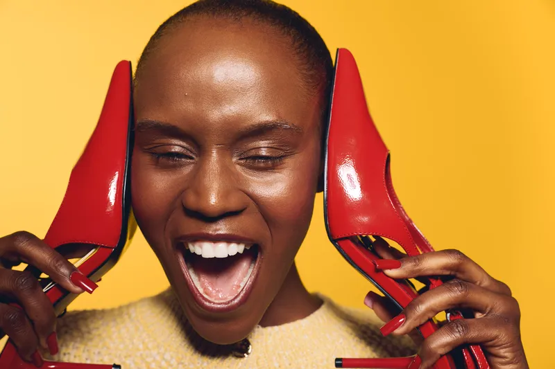 Happy black African woman with dark skin holds red high heels near her face, laughing with closed eyes. Bright yellow background and joyful mood create a vibrant, fashion concept.