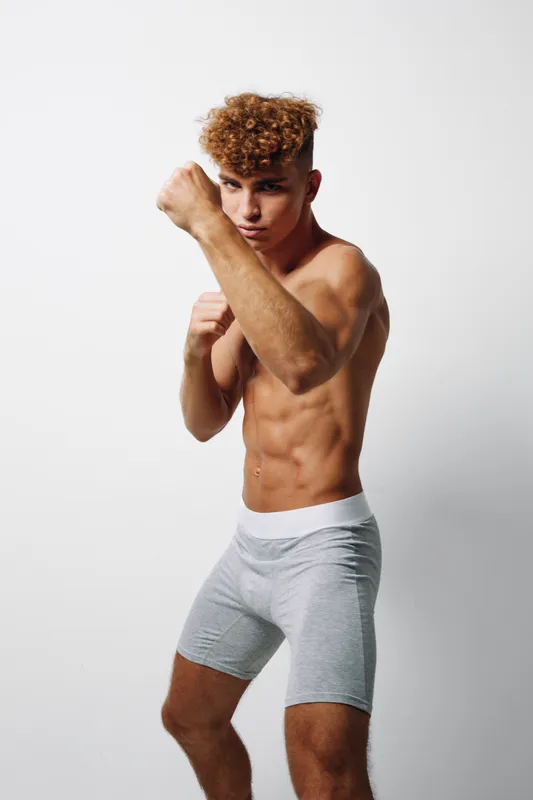 Athletic young man with fit muscular body in gray shorts practicing boxing stance white background focused confident strength and health lifestyle boxing training exercise.