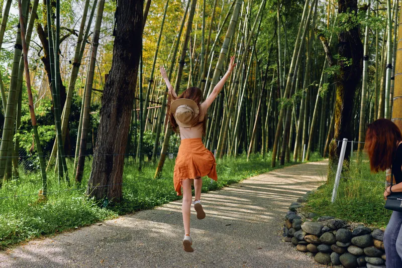 woman jump on a gravel path through a sunlit bamboo forest, orange skirt and straw hat visible as she raises her arms in carefree motion and joyful summer outdoor travel scene