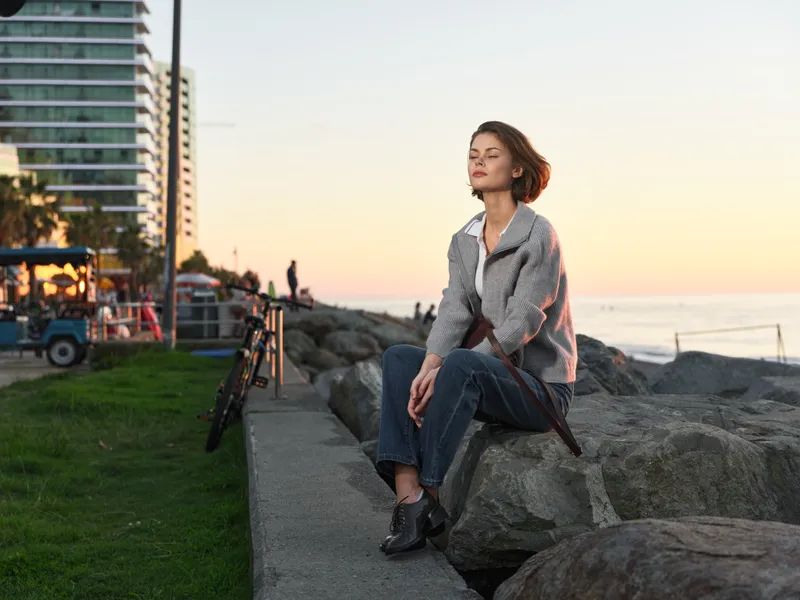 A thoughtful woman sits on a rocky shoreline at sunset, with distant city buildings and calm water behind her. She wears a blazer, jeans, and boots, looking contemplative and confident.