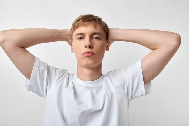 Man with arms behind head in a casual studio shot, white t shirt, relaxed expression, clean background, simple styling, youthfulness, confidence, everyday lifestyle, minimal and fresh.