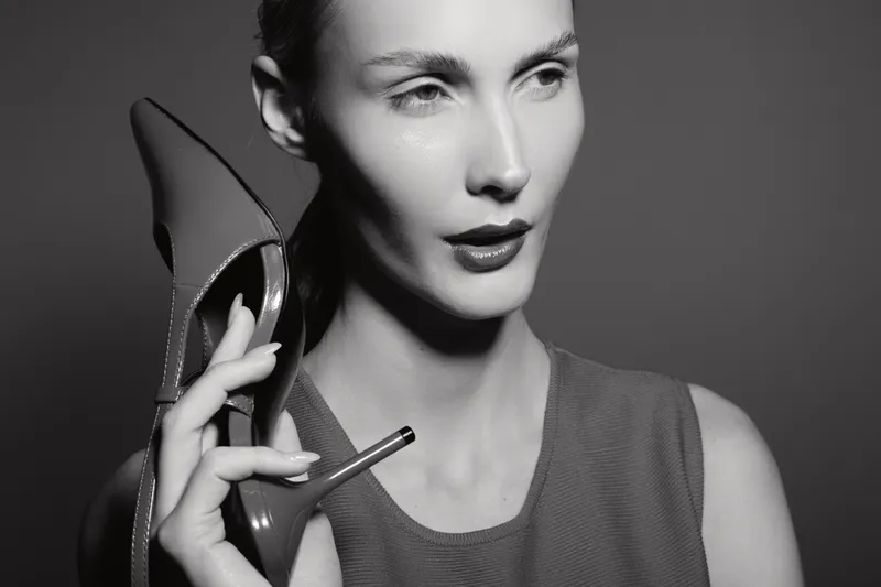Caucasian woman with fair skin holding high heel shoe near face in black and white portrait. Concept of fashion, elegance, and style in studio lighting.