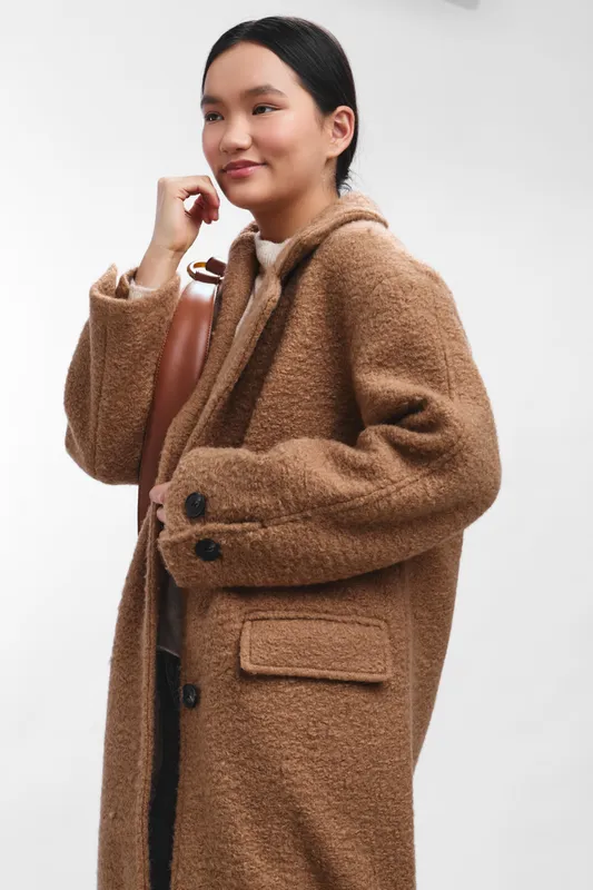 Asian woman wearing a warm wool coat stands in a clean studio, carrying a bag and looking ahead with calm confidence, conveying poised urban style.