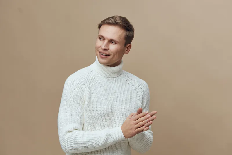 Smiling young man with short hair wearing a white knitted sweater gesturing with his hand, looking away, in a studio with beige background, casual fashion, positive expression, lifestyle concept.