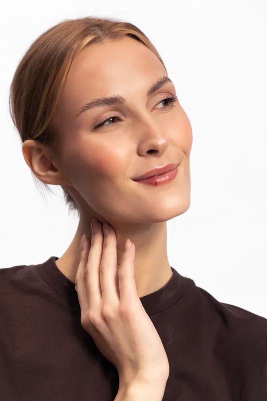 Beautiful young woman with clear skin and light brown hair touches her neck gently, looking away with a soft smile. Concept for skincare, beauty, natural, and wellness.