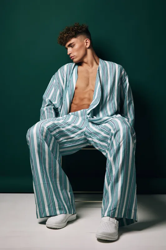 Young man with curly hair wearing striped loose pajamas sitting on chair against dark green background, casual relaxed pose with bare chest and white shoes in studio.