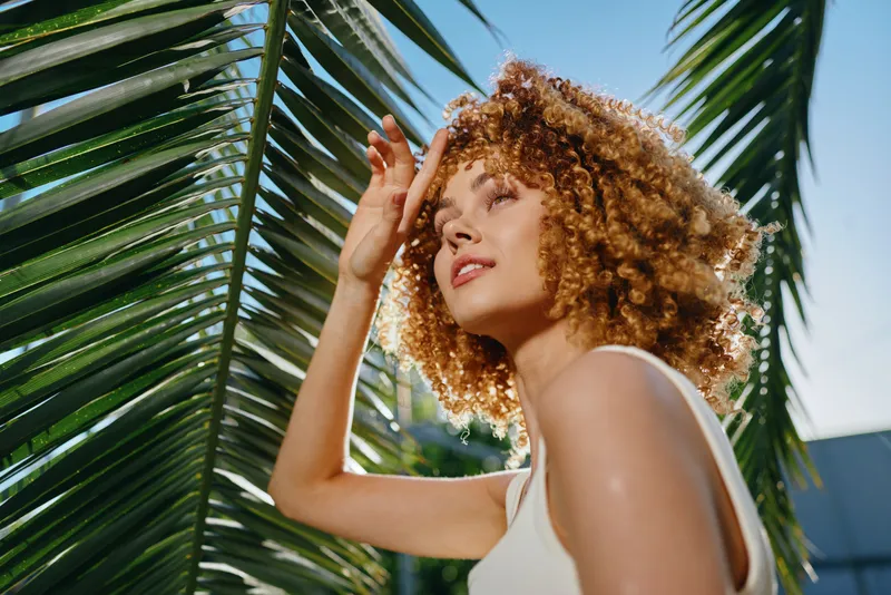 Curly woman enjoying tropical sunlight outdoors, surrounded by green palm leaves under a clear blue sky, radiating natural beauty and warmth.