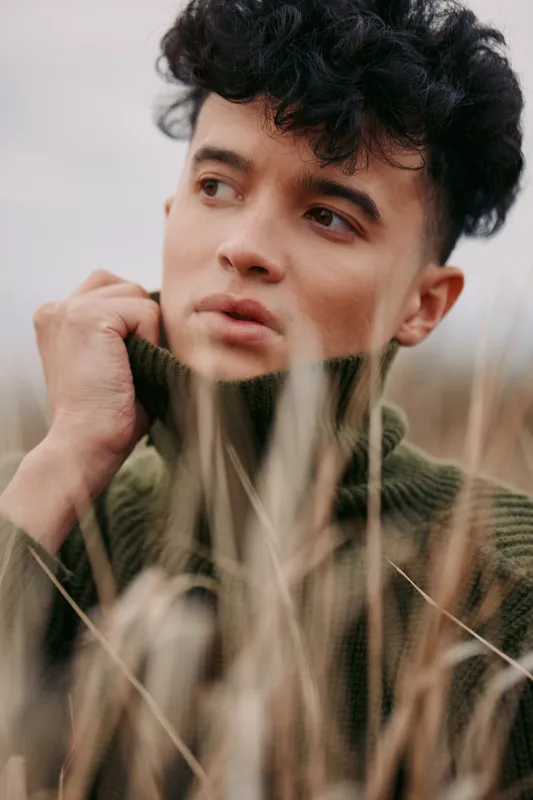 A candid portrait of a young man gazing away among tall grasses, wearing a chunky knit sweater. The image conveys genuine emotion, quiet focus, and authentic presence.