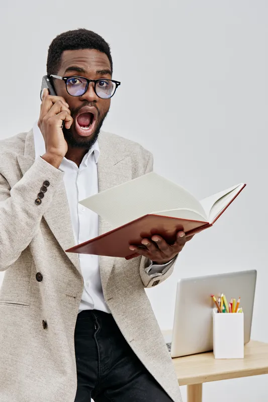 Engaged man with glasses talking on mobile phone while holding open book indoors, casual office background with stationery, surprised expression, diverse ethnicity, professional lifestyle concept