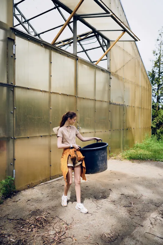 Girl teenager woman carrying a bucket near a greenhouse, gardening activity with plants and seedlings, casual clothing and sneakers on a sunny outdoor pathway by glass structure