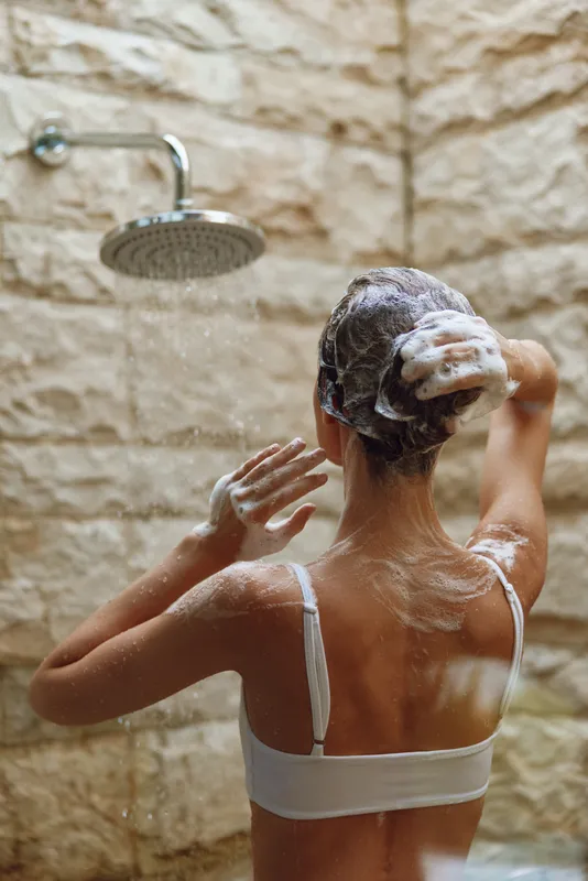 Woman washing hair in shower with white soap foam on short dark hair, beige tiled background, personal hygiene and skincare routine concept.