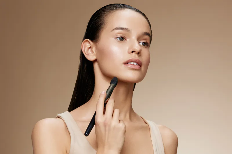 A young woman applying makeup with a makeup brush, showcasing a natural beauty look, soft skin tones, and a neutral background for beauty and skincare concepts