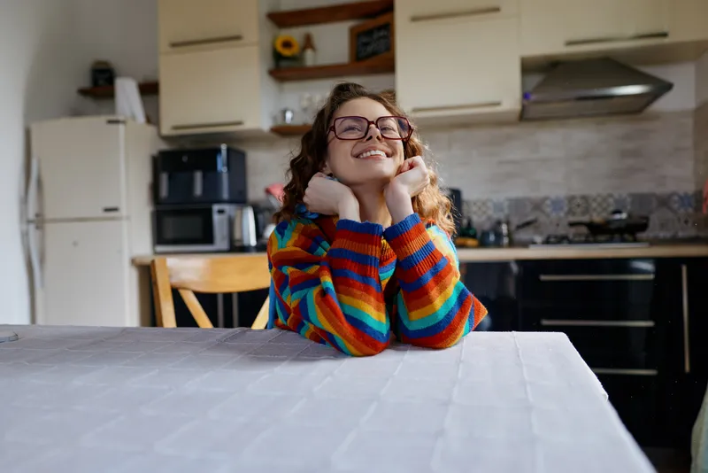 Smiling woman sits at a table in a bright kitchen, enjoying a cozy home moment in a colorful striped sweater, radiating warm happiness and relaxed lifestyle at home.