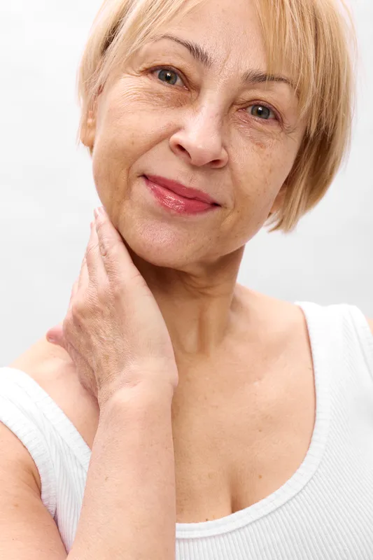 Elderly woman with short blond hair rests her hand on chin, thoughtful expression, bright white background, concept of aging, mature, stress, self care