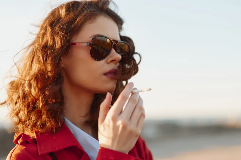 Young woman with wavy hair in a red coat wears sunglasses and smokes on a sunny beach, projecting confidence, a cool, relaxed mood, and golden hour breeze.