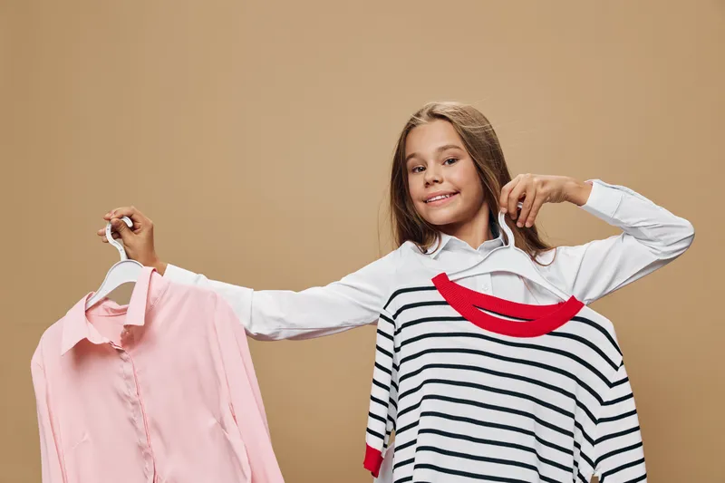 Girl choosing between two shirts holding one in each hand with smile on beige background. Young teenager comparing clothing for casual style and fashion decision indoors.