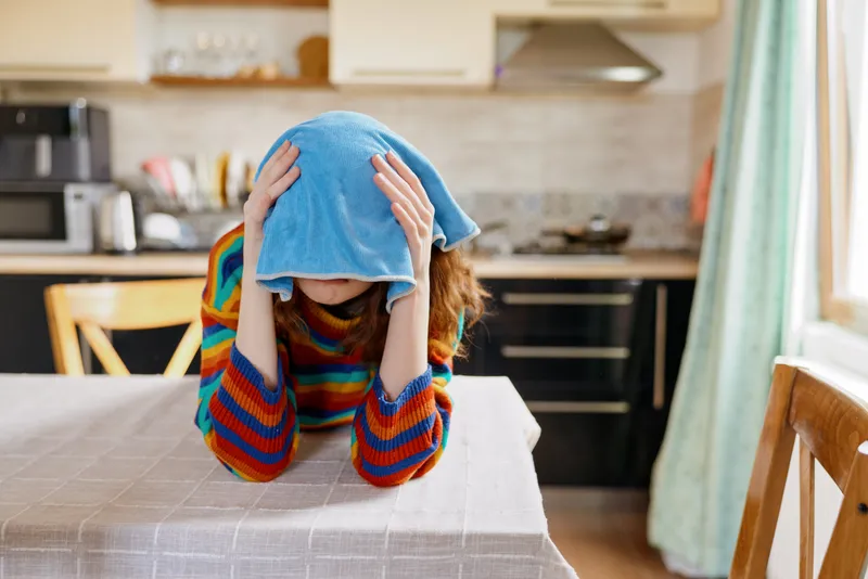 A distressed woman sits at a kitchen table, head covered by a blue towel, conveying fatigue and overwhelm in a cozy home kitchen. Concept for stress, burnout, and mental health in daily life.