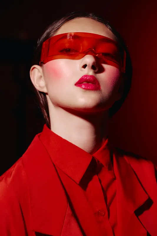 Styled young woman with glossy red lips and enhanced blush wears red translucent futuristic glasses. She poses confidently in a monochrome red outfit under dark background. Beauty fashion glow.