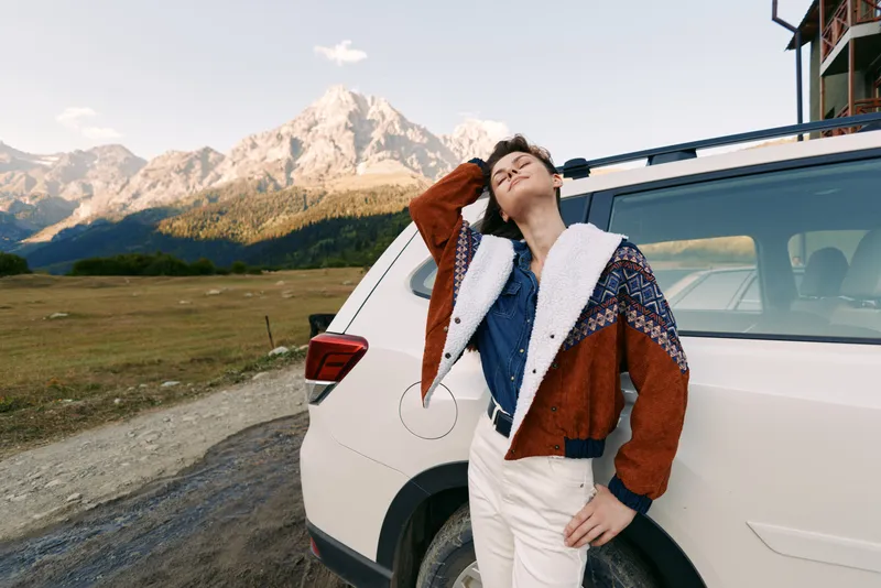 Woman, car, mountains, headphones, travel, roadtrip, nature, relaxation: Young woman leans against white SUV on a mountain road, wearing headphones and warm jacket, eyes closed enjoying music.