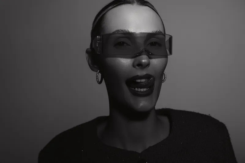 Portrait of a light skinned Caucasian woman wearing futuristic visor glasses and dark lipstick, posing with tongue out in studio lighting on plain grey background.