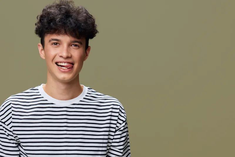 smiling man with curly hair wearing striped shirt sticks out tongue in front of plain olive background, expressing playful and fun mood