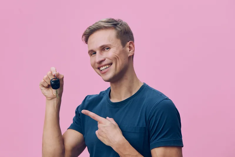 Smiling man holding a small black object, pointing at it with his finger, wearing casual blue t-shirt, energetic expression, studio shot, pink background, concept of curiosity and discovery.