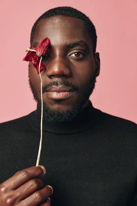 African man with dark skin holding red flower near face in creative conceptual visual style. Portrait of black male model with beard wearing black turtleneck on pink background for artistic