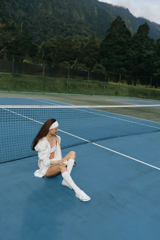 Young female tennis player in sporty outfit sitting on the court, expressing relaxation and concentration outdoors in nature setting