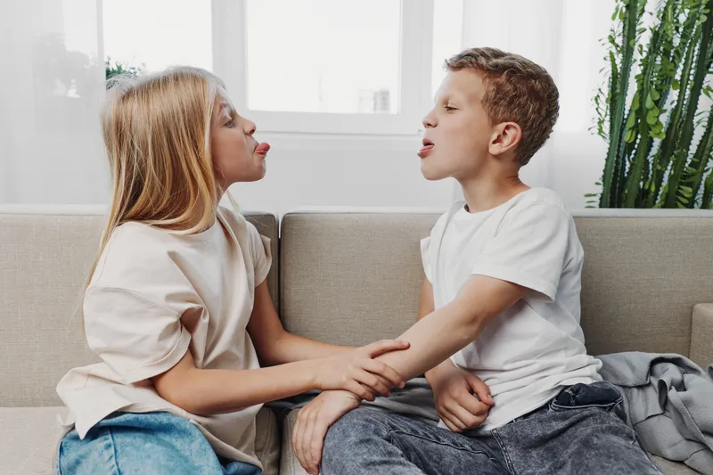 Two children sitting on a sofa making funny faces and holding hands in a bright living room. Concept of sibling fun, family bonding, and playful interaction at home.