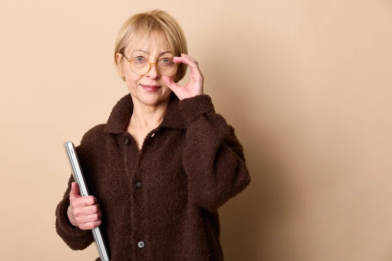 Elderly woman with glasses holds a smartphone, touching her ear, looking concerned on a beige background, concept for daily communication troubles, aging, loneliness now