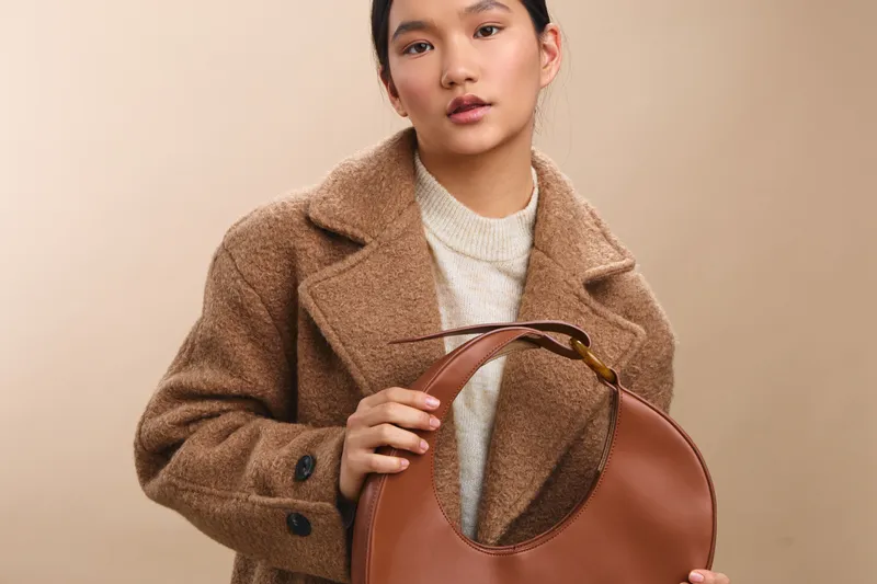 asian woman wearing a warm beige coat gazes softly at the camera while gripping a brown handbag, presenting a poised and stylish autumn look in a calm studio setting.