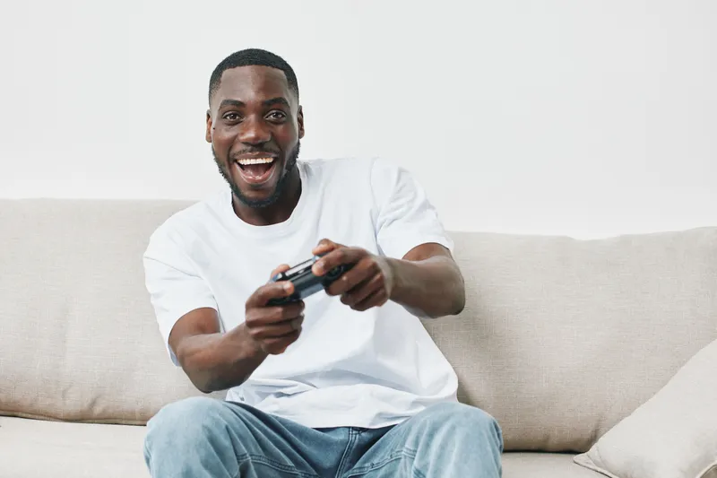 Happy young African American man playing video games at home, showcasing excitement and joy in a casual setting with a light background