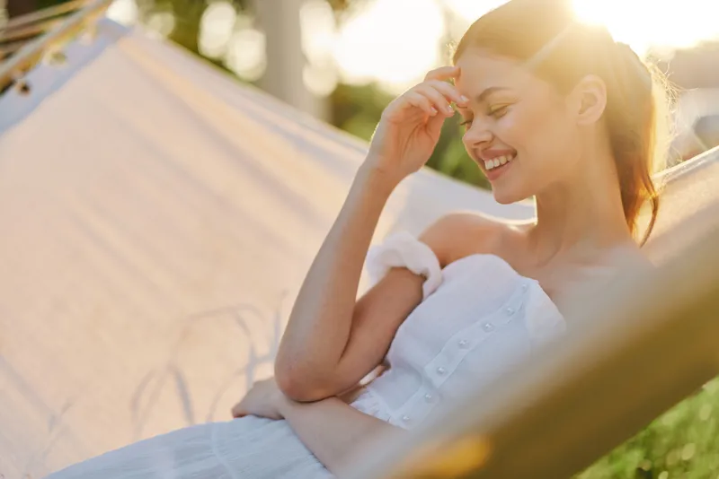 Young woman relaxing in a hammock, smiling and enjoying the sunshine in a white dress, surrounded by greenery, conveying a sense of peace and joy
