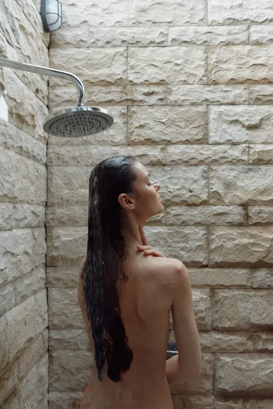 naked woman under rain shower with wet hair, indoor stone wall, serene expression, natural light, calm atmosphere, intimate moment captured