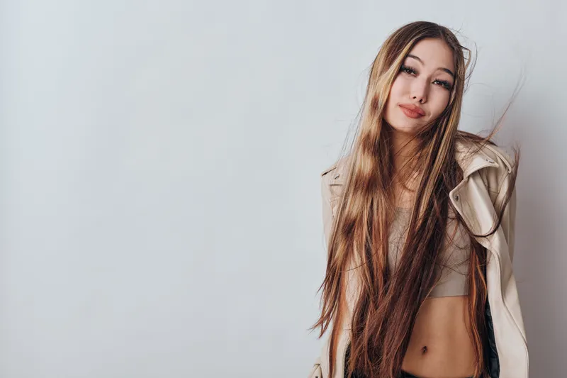 Young woman with long hair posing in beige coat and crop top against light gray background. Studio portrait of attractive female with casual confident look and natural makeup.