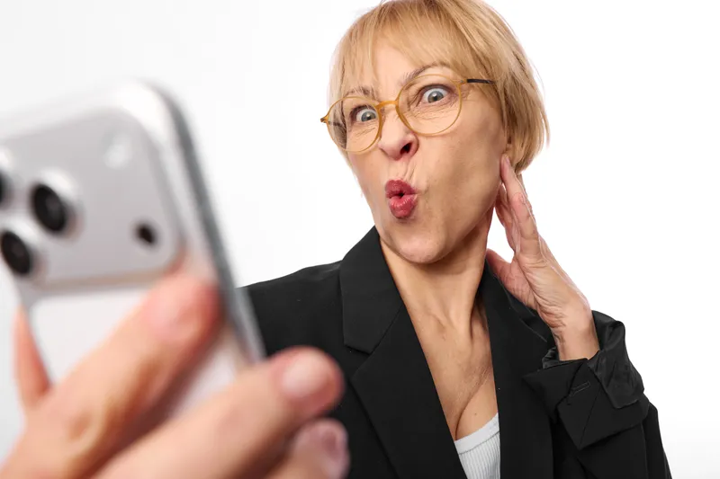 Surprised senior woman with glasses takes a selfie on smartphone, face astonished with open mouth, indoor portrait on white background, communication and technology