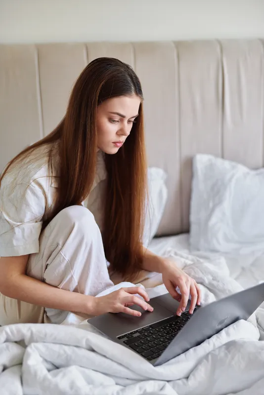 Woman on bed typing on laptop at home in casual pajamas, remote work or study scene, focused portrait with long hair and cozy bedding, lifestyle image for online learning and freelance work