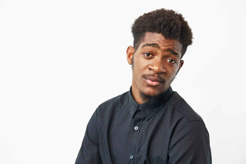 A thoughtful black man in a black shirt, displaying a pensive expression and relaxed posture. His unique style adds character to the composition, capturing a moment of reflection.