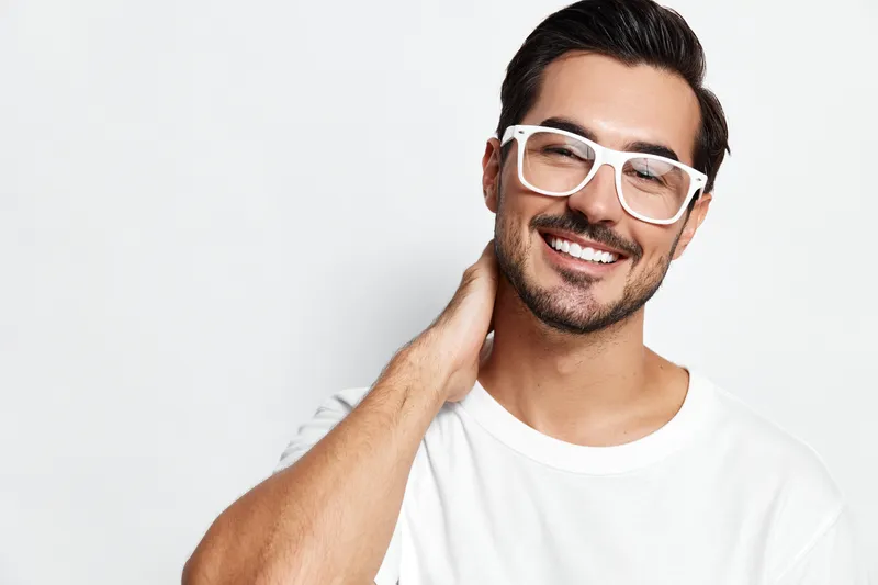 Smiling man with glasses poses confidently while touching the back of his neck, showcasing a charming expression against a plain light background.