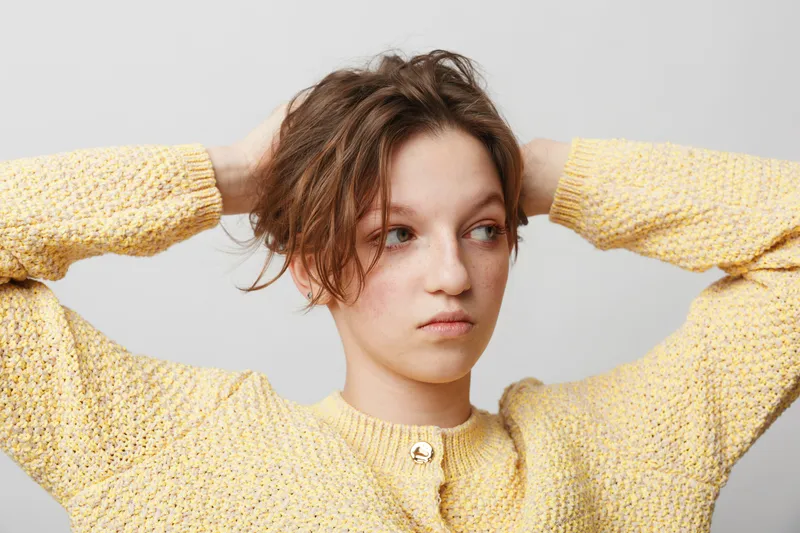 Young woman in yellow knitted sweater looks away with hands behind head. Casual style, natural light, and neutral background create a relaxed and thoughtful mood.