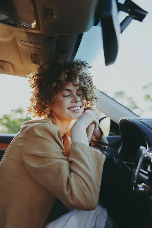A young woman sits in a car, smiling with eyes closed, hands clasped near chin. Sunlight and retro film tones lend a warm, nostalgic lifestyle vibe, capturing calm, intimate travel mood.