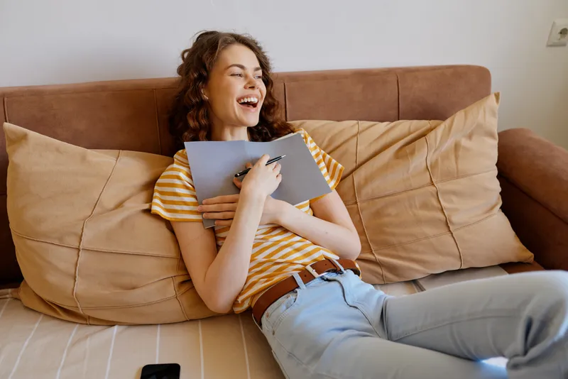 Smiling woman with curly hair sits on a sofa, writing in a notebook. Casual home setting with warm lighting, relaxed mood, and everyday creativity. Bright cozy scene conveys comfort.