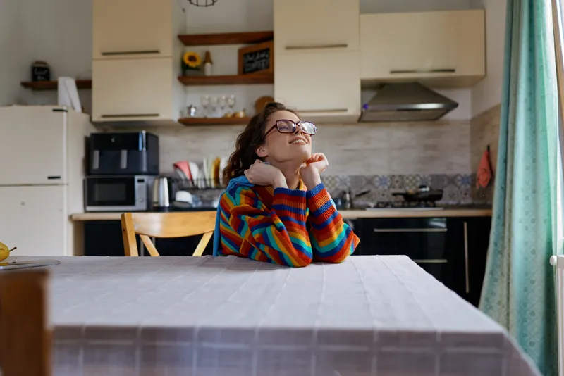Cheerful woman indoors at a sunlit kitchen table, resting chin on hands, colorful striped sweater, relaxed lifestyle moment, warm natural tones, cozy home ambience