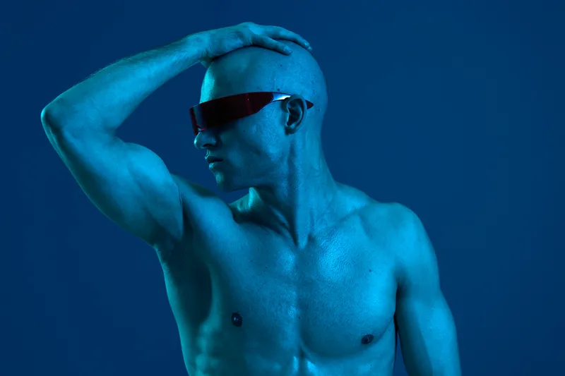Athletic man striking a pose wearing sleek sunglasses, showcasing a strong physique against a vibrant blue background. This image captures confidence and modernity.