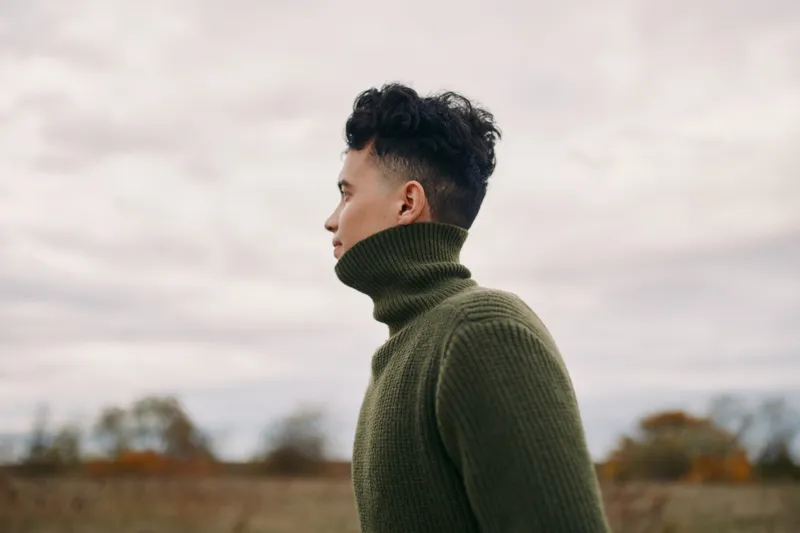 Profile portrait of a young man in a thick turtleneck sweater standing outdoors under a muted sky, conveying raw authenticity and quiet credibility through a contemplative, unposed lifestyle moment.