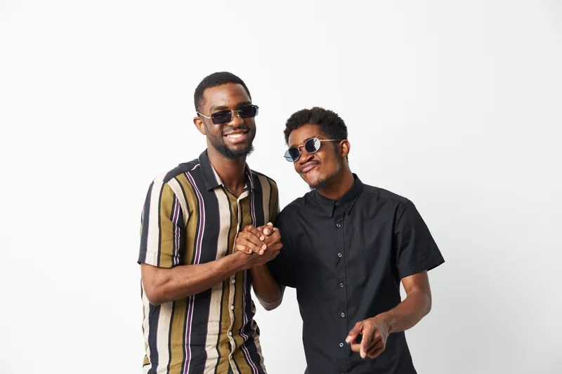 Two smiling African American men share a joyful moment together, dressed in stylish attire with sunglasses, radiating happiness and friendship in a bright setting.