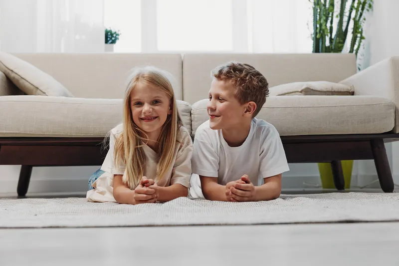 Smiling boy and girl lying on carpet in bright living room with modern sofa and plants. Children wearing casual white clothes enjoy relaxed moment in cozy home interior. Concept of family and