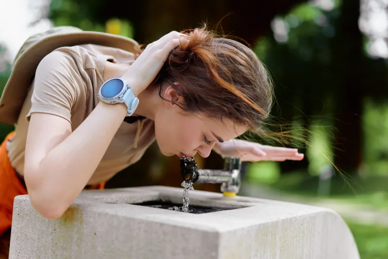 Woman drinking water from a public fountain outdoors in a park, leaning over concrete basin with backpack and smartwatch, casual summer scene and refreshing moment